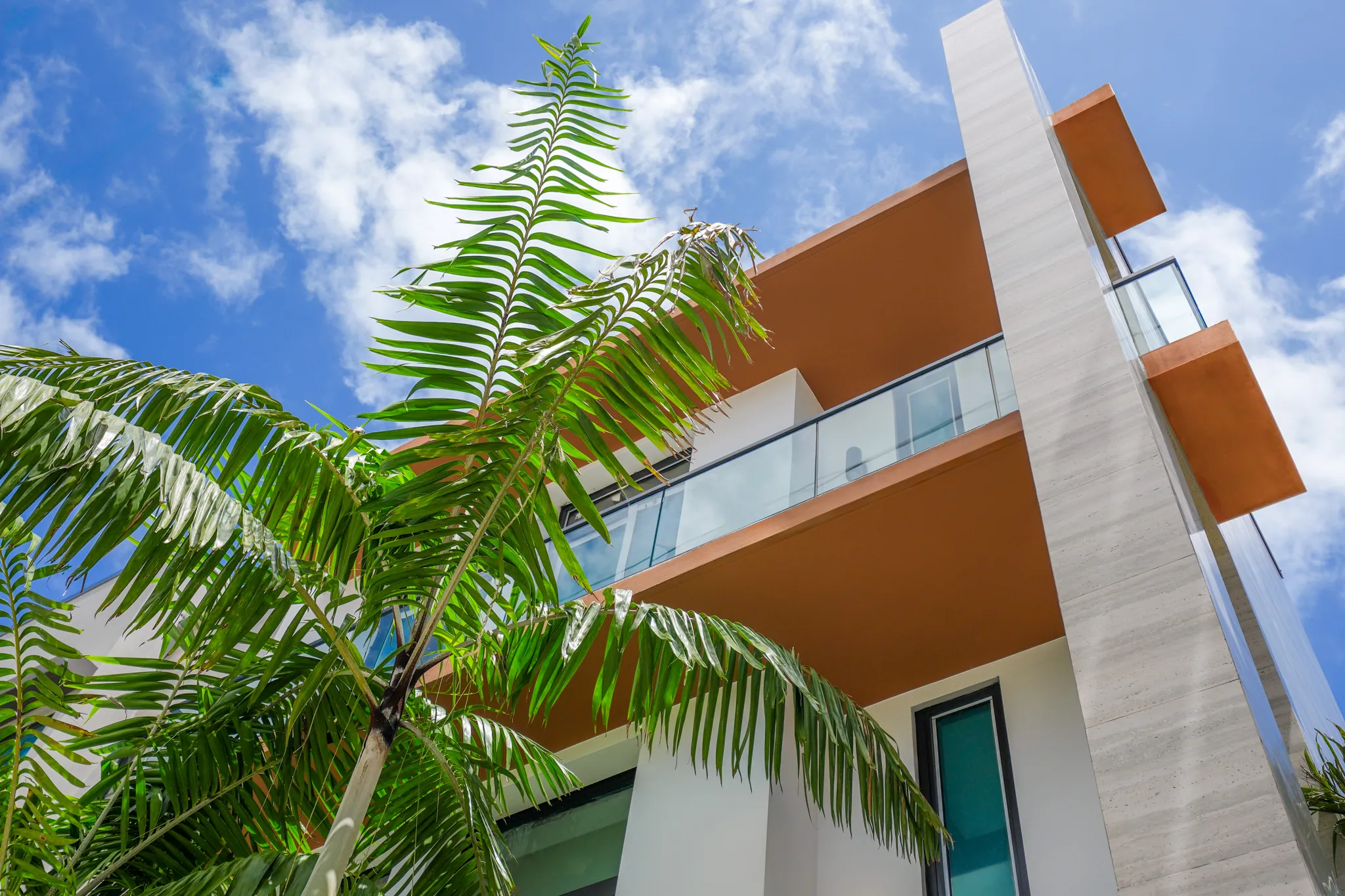 Upward view of cantilevered orange-trimmed balcony framed by palm fronds against partly cloudy sky