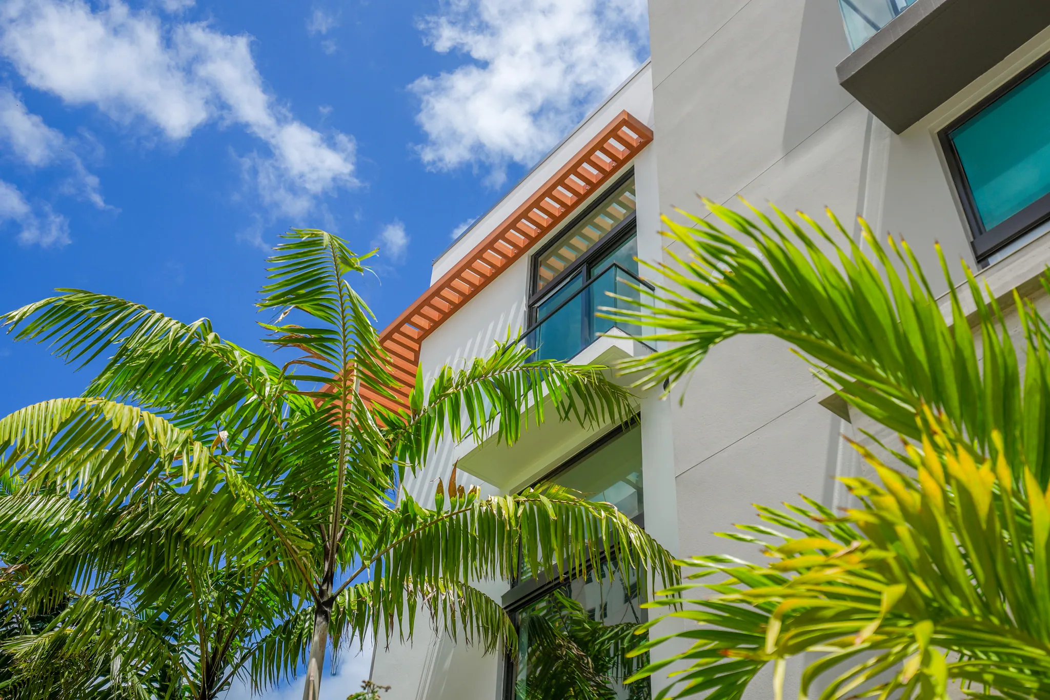 Looking up at white townhome facade with wood-slat overhang and palm trees in foreground
