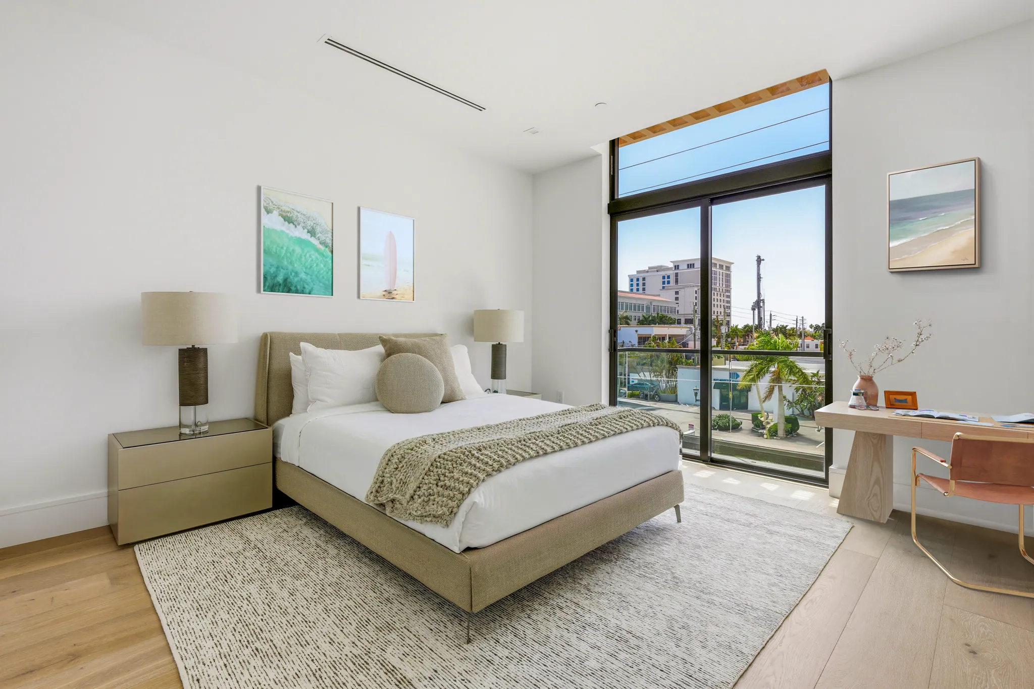Secondary bedroom with upholstered bed, work desk, and balcony overlooking palm-lined skyline