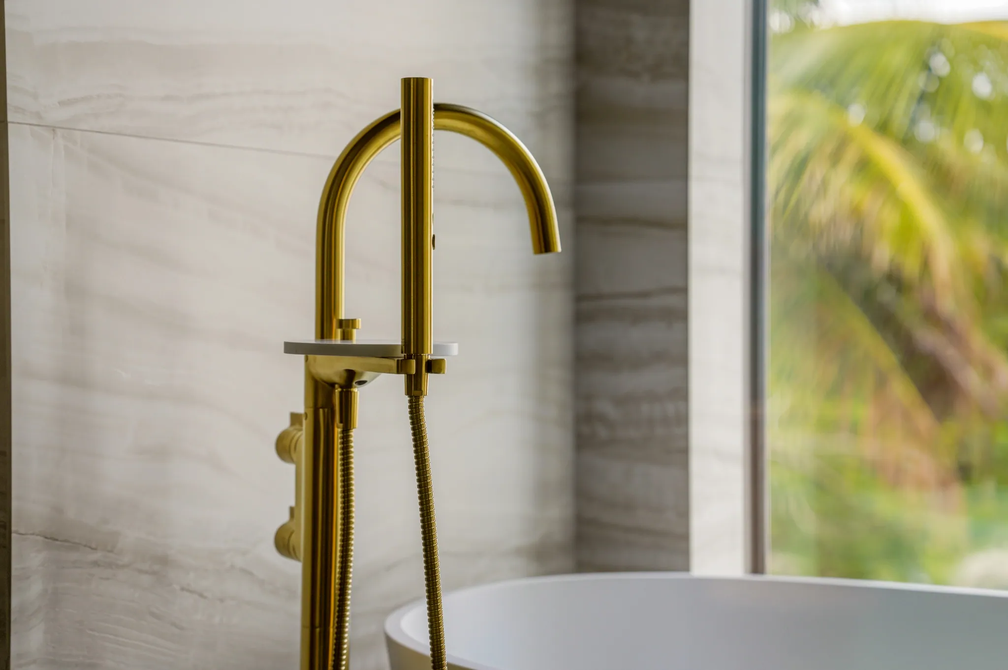 Floor-mounted brass tub filler beside freestanding bath with palm view